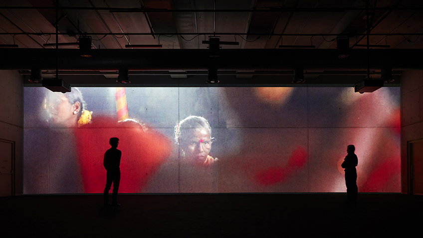 Two people stand in a dark room watching a large-scale film of a Indian spiritual festival Dhalo