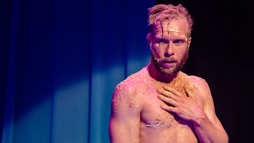 Joel Bray on stage shirtless against a blue backdrop.