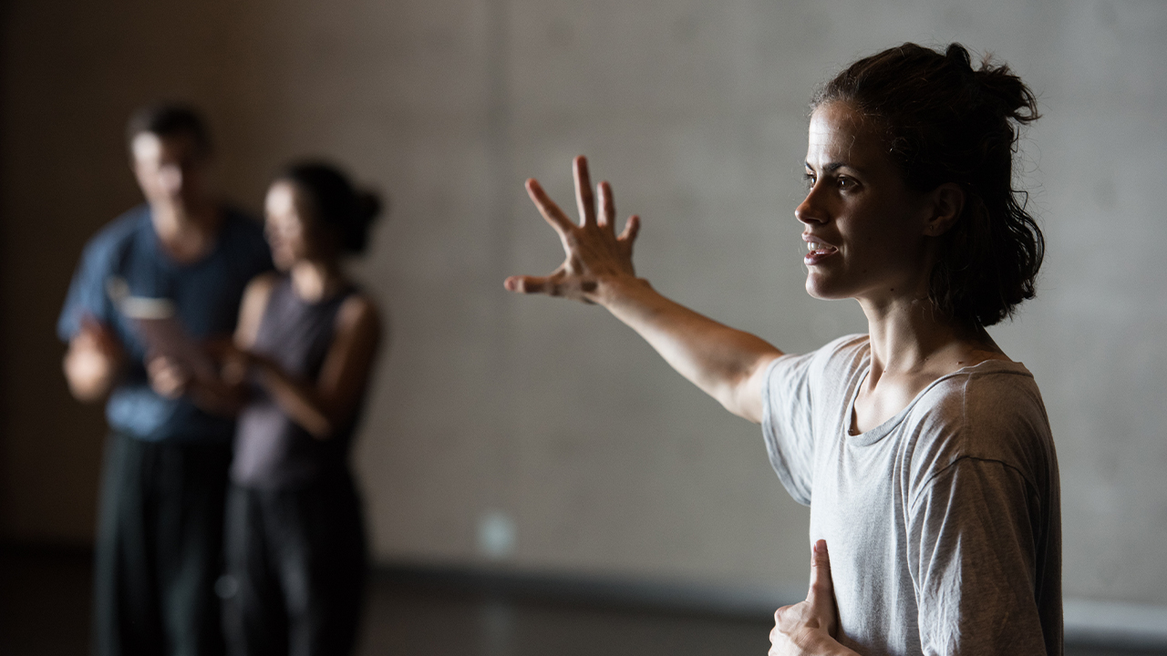 A person stands with their right hand stretched out. She's in a masterclass for Force Majeure. There are two other people blurred in the background.