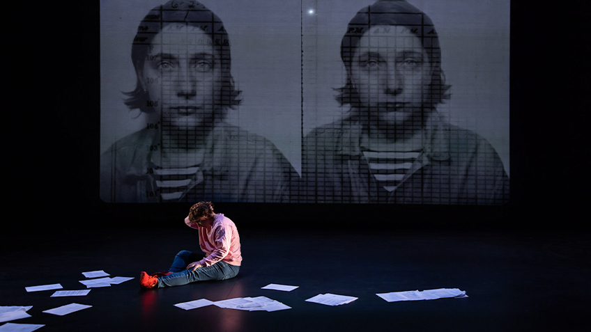 Kaz Therese sits on the ground with papers strewn on the floor. There is a headshot of someone projected twice behind them.