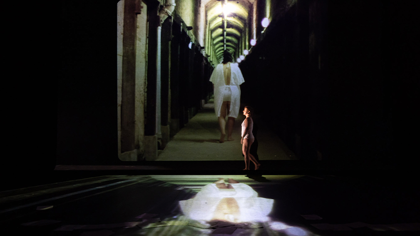 Kaz Theres walks across stage with a projection of them in a hospital gown in the background.