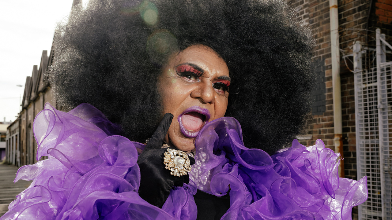Nana Miss Koori wears a black afro and purple ruffle coat with black gloves and a huge statement ring.