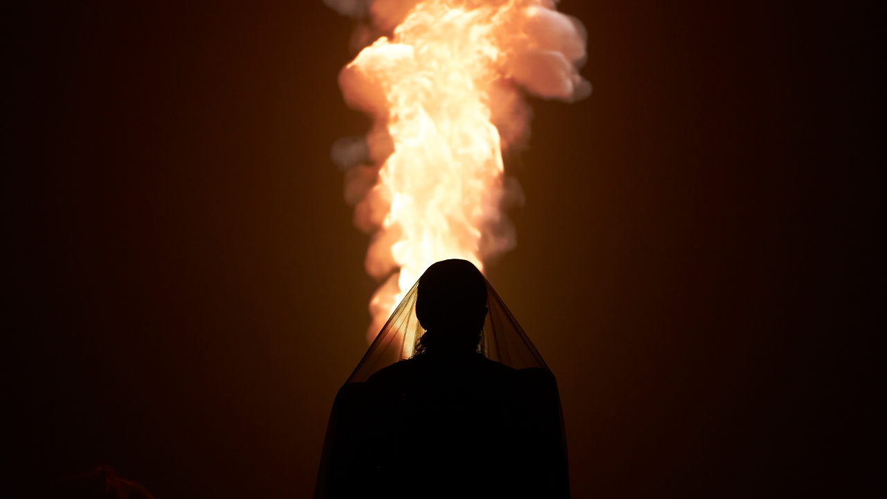 A veiled figure stands in front of what looks like fire.